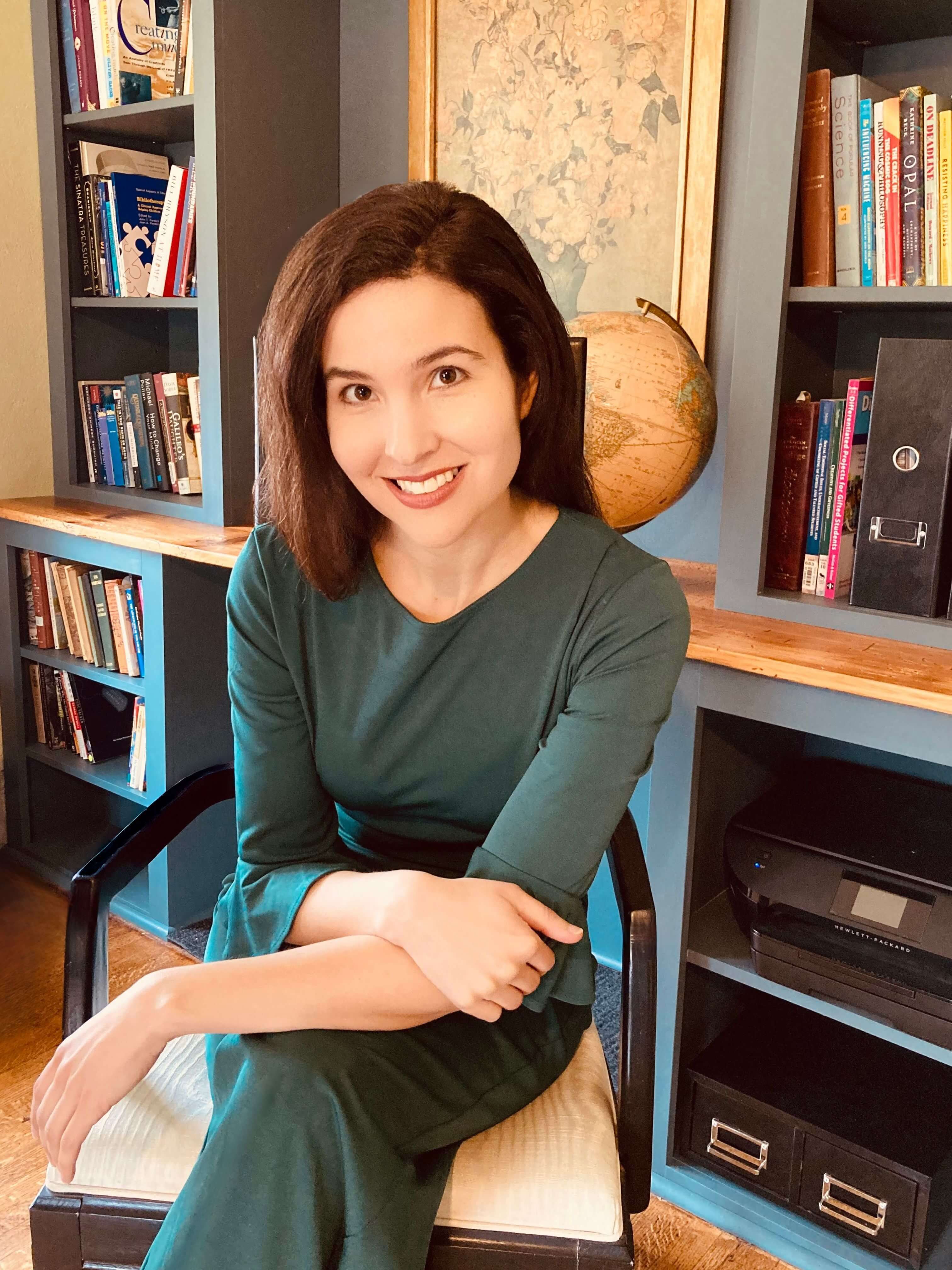 Michelle Ptacek, seated, with long brown hair and a green sweater smiles in front of a framed floral picture and bookcase.