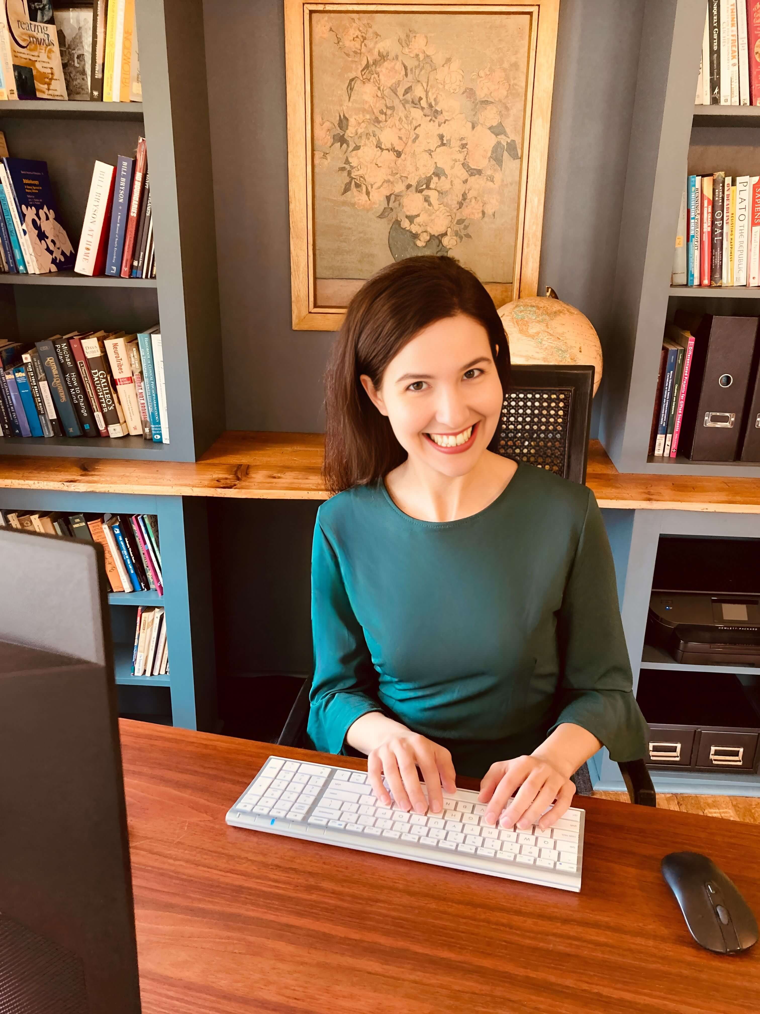 Michelle Ptacek in a green blouse smiles in front of a framed floral picture and bookcase.