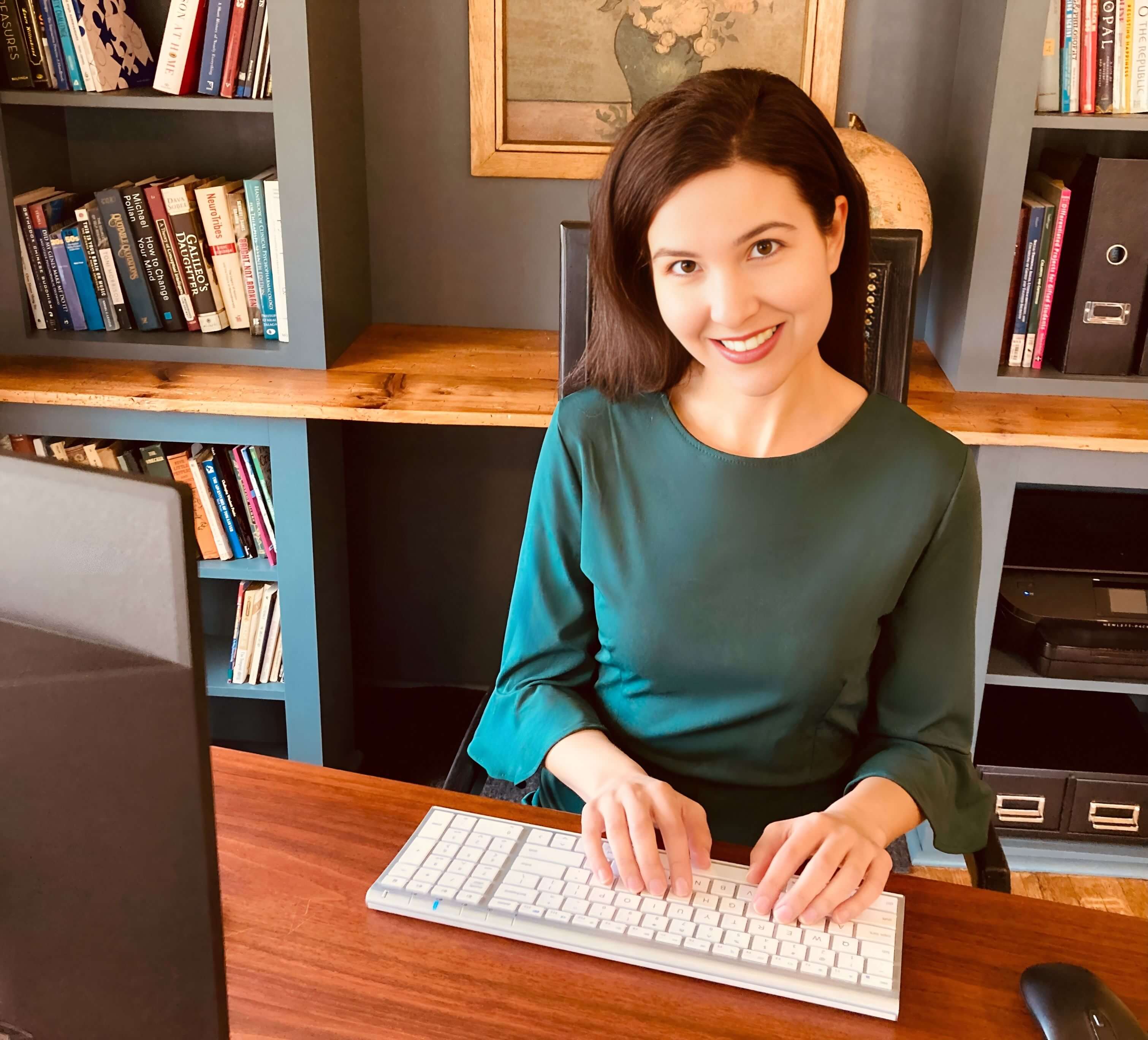 Michelle Ptacek in a green sweater smiles while typing on white keyboard in front of a framed floral picture and bookcase.