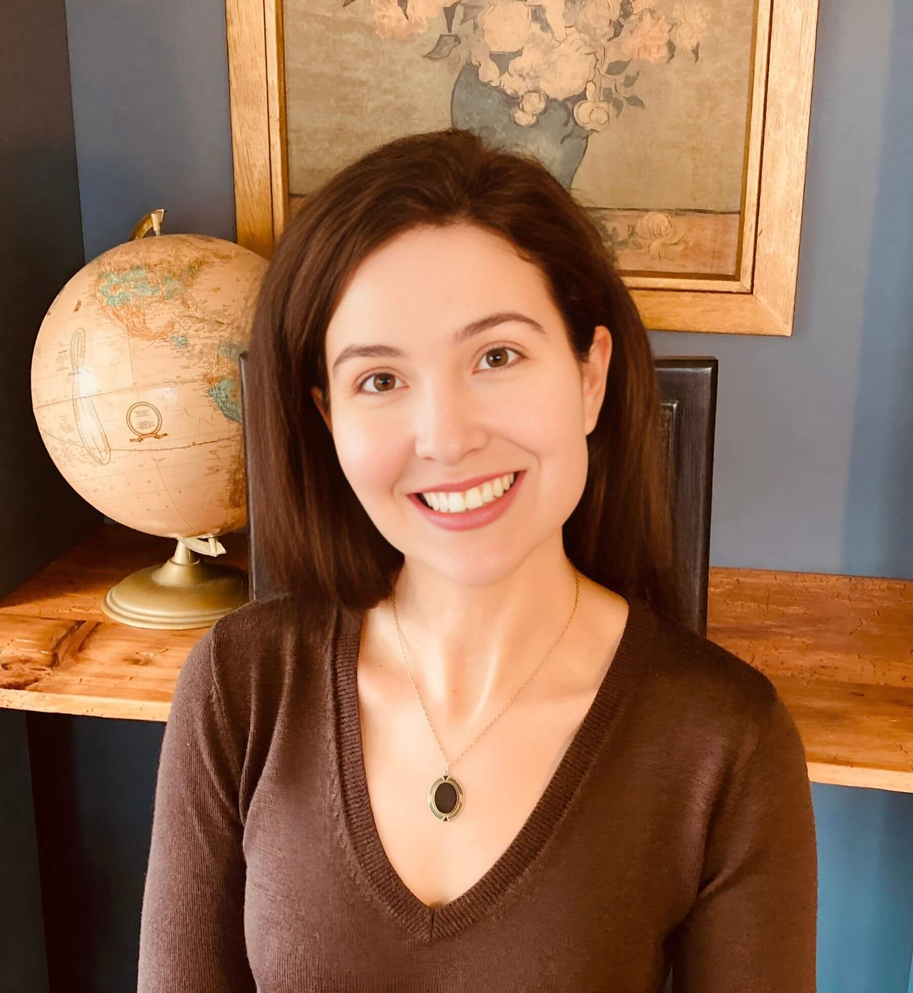 Michelle Ptacek wearing a brown sweater smiles in front of a framed floral picture and globe.