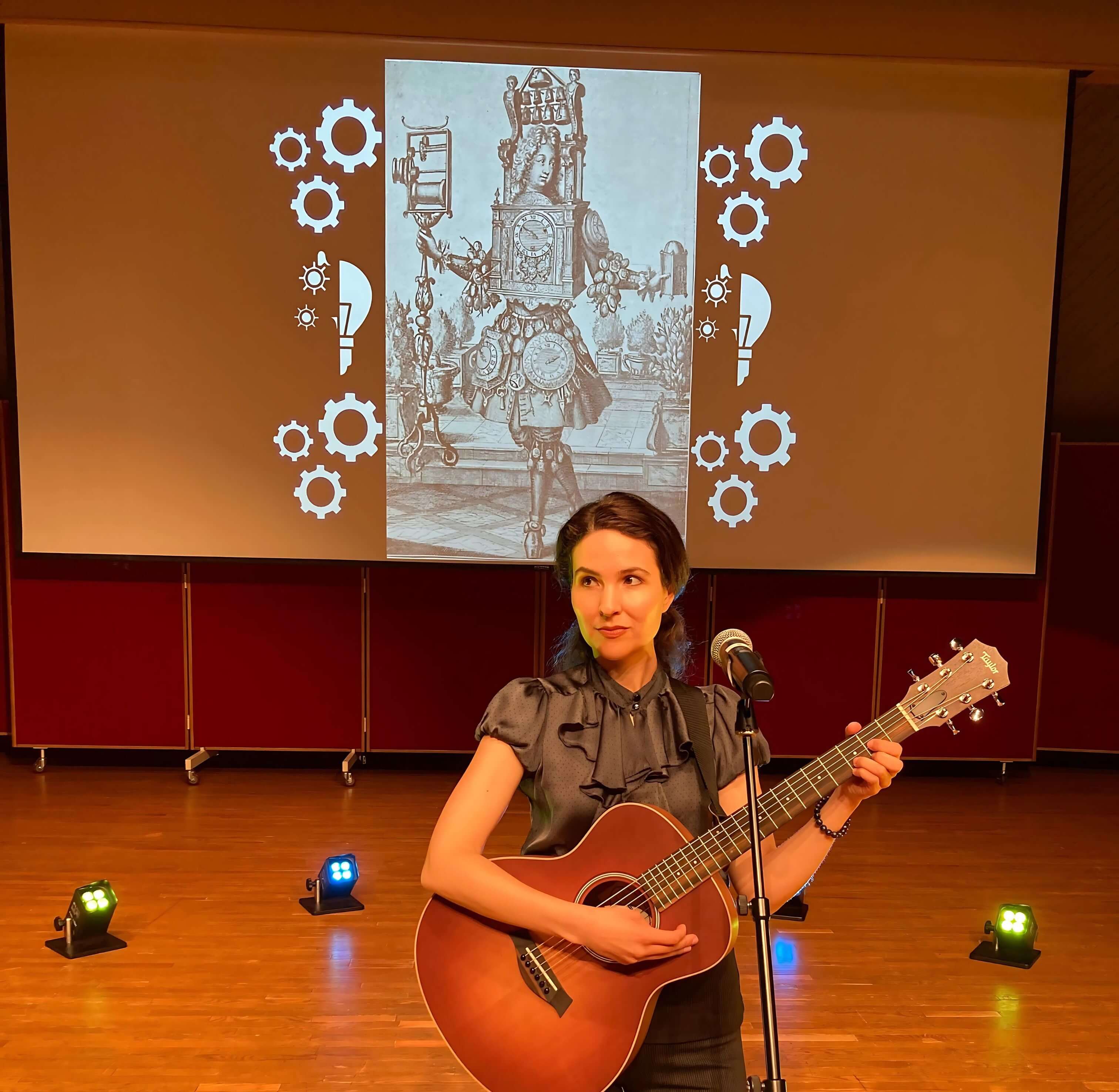 Michelle Ptacek stands on a stage, playing an acoustic guitar with a microphone. Behind her is a large projection screen displaying a vintage illustration with gears around the image.