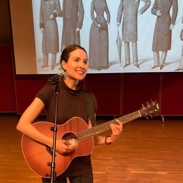 Michelle Ptacek stands on a stage, playing an acoustic guitar with a microphone. Behind her is a large projection screen displaying a vintage illustration of six individuals.