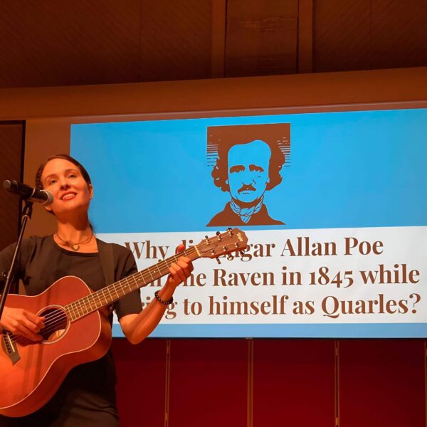 Michelle Ptacek playing an acoustic guitar with a microphone. Behind her is a large projection screen with a blue background displaying a illustration of Edgar Allan Poe.