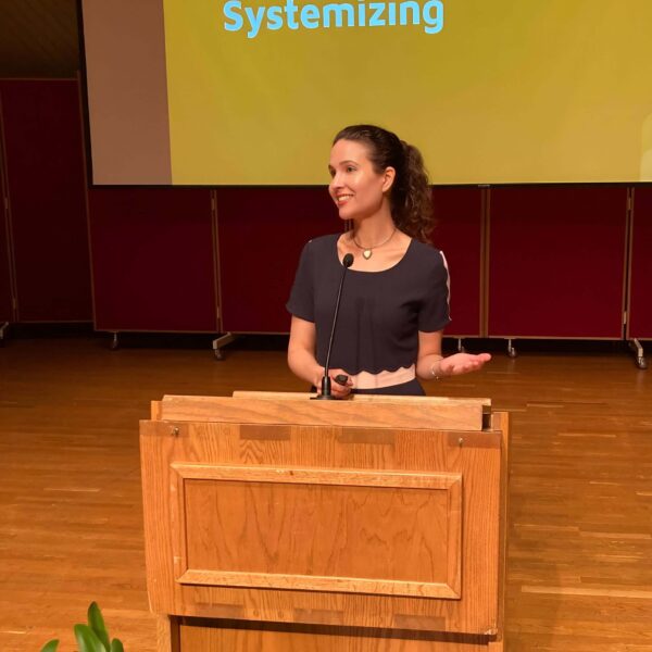 Michelle Ptacek in a black top stands on a stage behind a podium. Behind her is a large projection screen with a presentation slide saying "Empathizing and Systemizing".