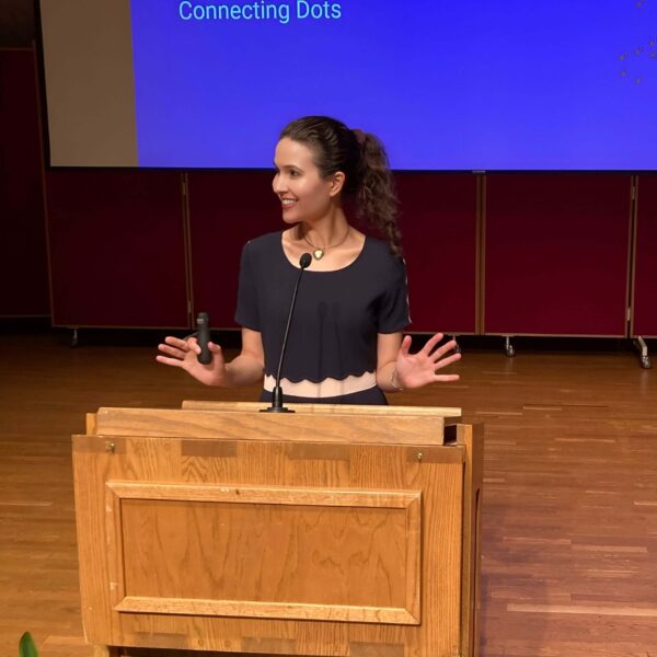 Michelle Ptacek in a black top stands on a stage behind a podium. Behind her is a large projection screen with a presentation slide saying "Analogous Thinking - Collecting Dots, Connecting Dots".