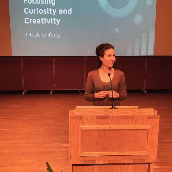 Michelle Ptacek stands on a stage behind a podium. Behind her is a large projection screen with a presentation slide saying "Focusing Curiosity and Creativity + task-shifting".