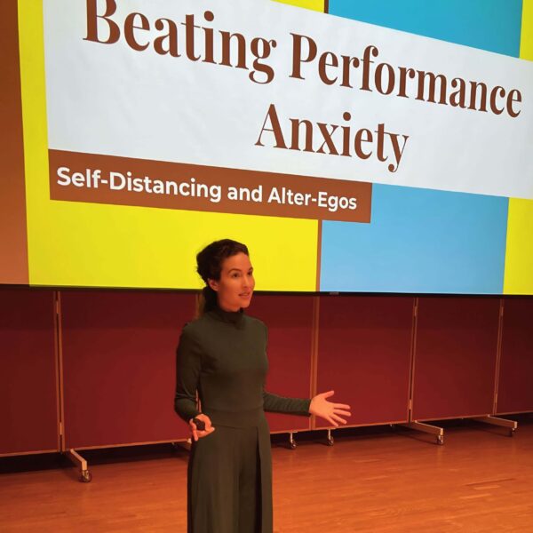 Michelle Ptacek wearing a green dress standing in front of a projection screen with a presentation slide saying "Beating Performance Anxiety - Self-Distancing and Alter-Egos".