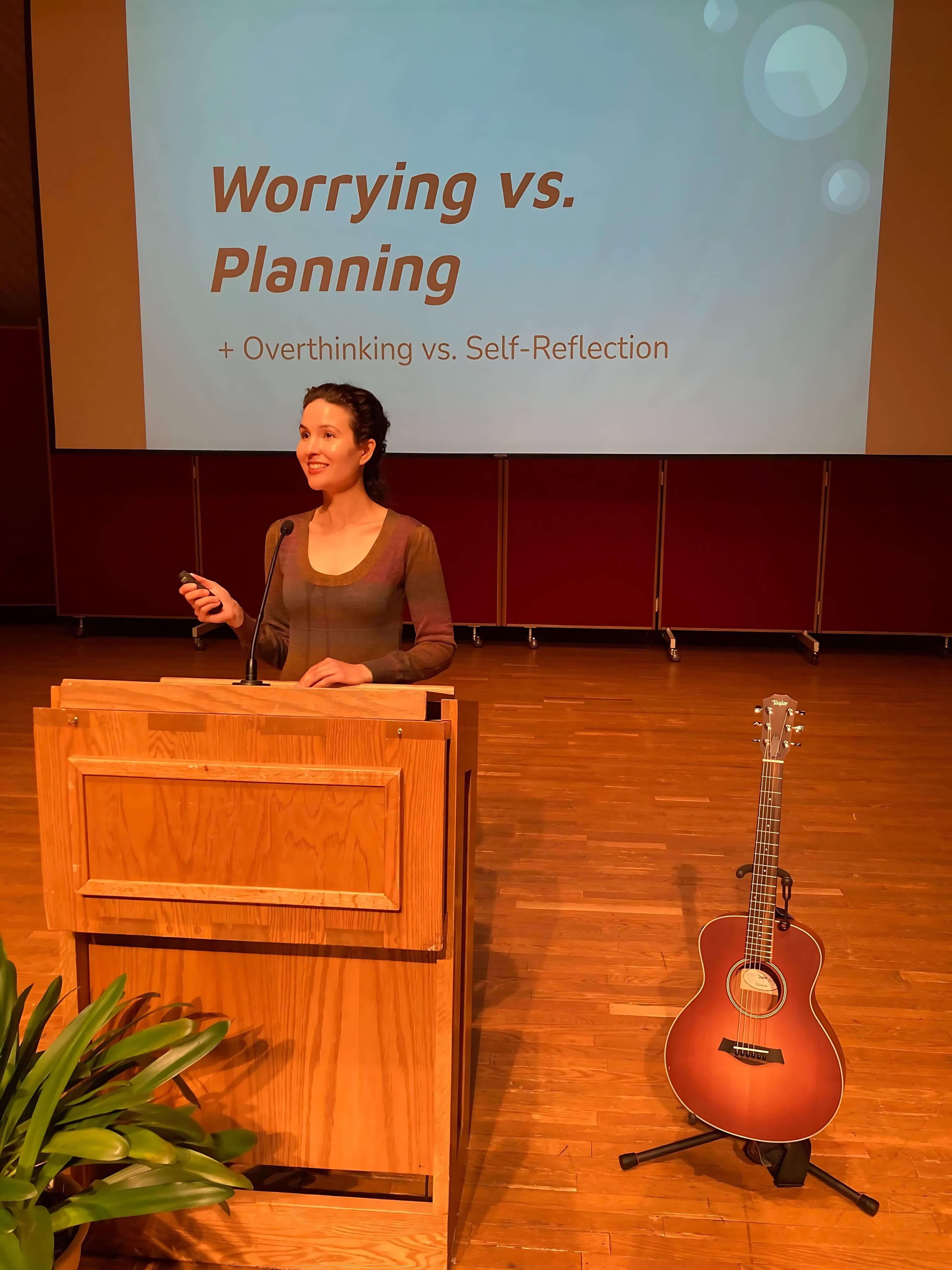 Michelle Ptacek stands on a stage behind a podium with an acoustic guitar on the floor next to her. Behind her is a large projection screen with a presentation slide saying "Worrying vs. Planning + Overthinking vs Self-Reflection".