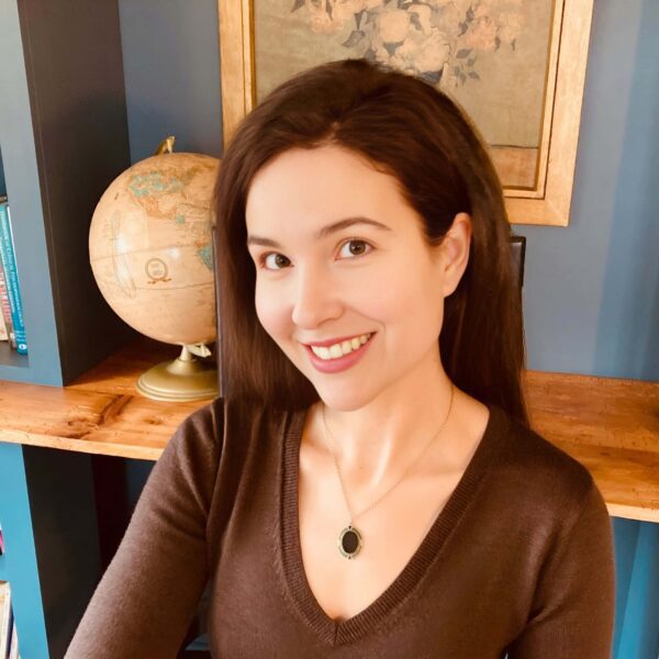 Michelle Ptacek in a brown sweater smiles in front of a framed floral picture and globe. Her head is slightly tilted away from camera.