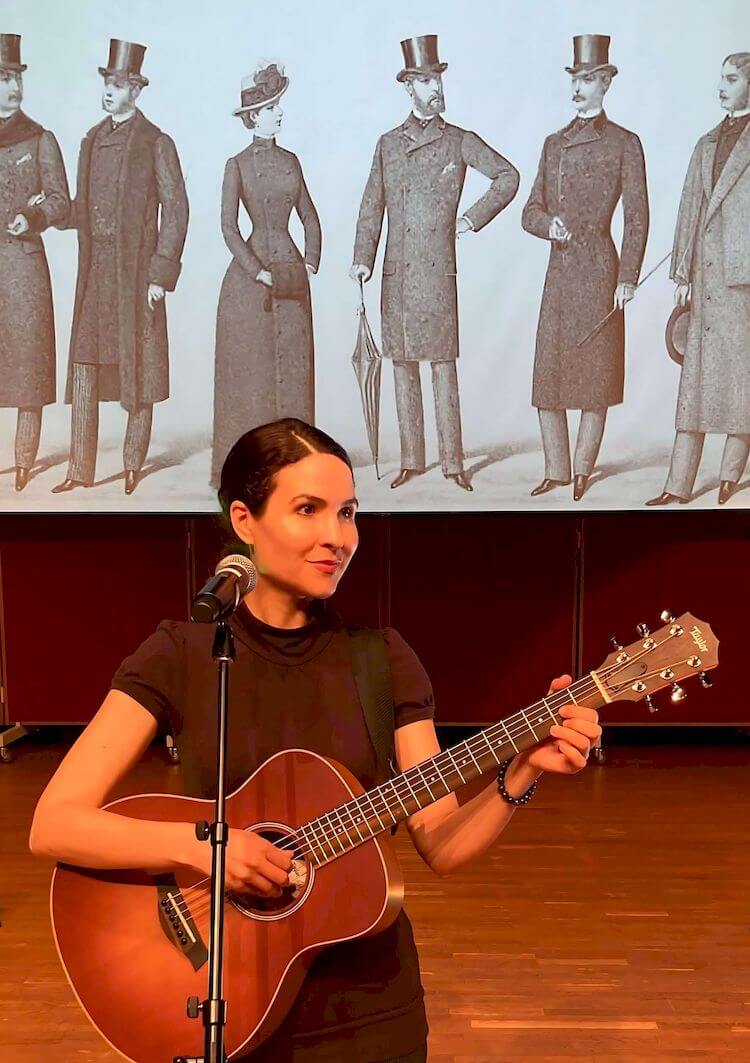 Michelle Ptacek stands on a stage, playing an acoustic guitar with a microphone. Behind her is a large projection screen displaying a vintage illustration of six individuals.