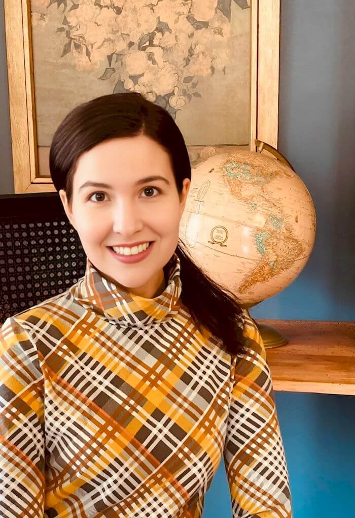 Michelle Ptacek in a patterned shirt smiles in front of a globe and a framed floral picture.